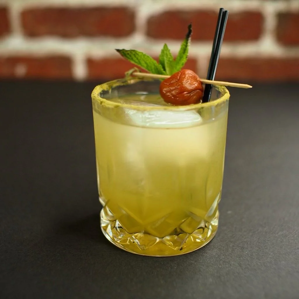 A chilled yellow cocktail garnished with a green leaf and cherry on Kinda’s wooden table.